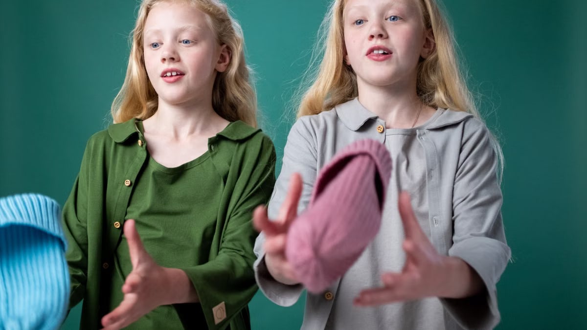 Twin girls wearing casual outfits, playfully tossing hats indoors against a green background.